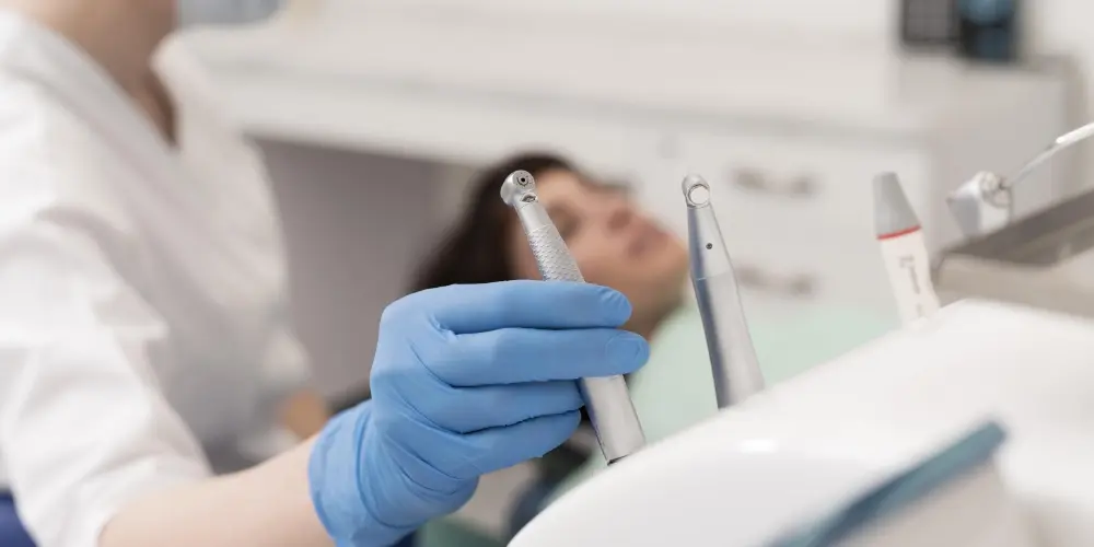 Dentista con guanti blu seleziona uno strumento odontoiatrico da un vassoio, mentre una paziente attende sullo sfondo nella poltrona dello studio.