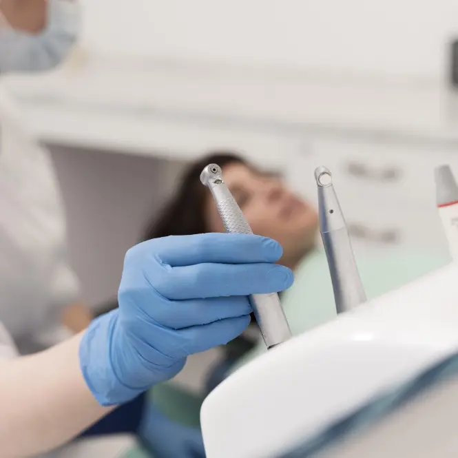 Dentista con guanti blu seleziona uno strumento odontoiatrico da un vassoio, mentre una paziente attende sullo sfondo nella poltrona dello studio.